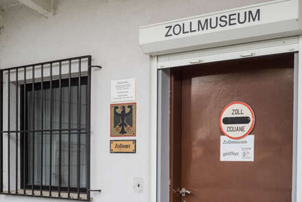 Eine braune Tür über der "Zollmuseum" steht. Daneben ein vergittertes Fenster.