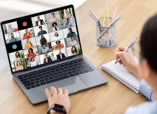 Eine Frau sitzt an einem Schreibtisch. Vor ihr steht ein geöffneter Laptop. Auf dem Bildschirm läuft eine Videokonferenz mit mehreren Teilnehmern.