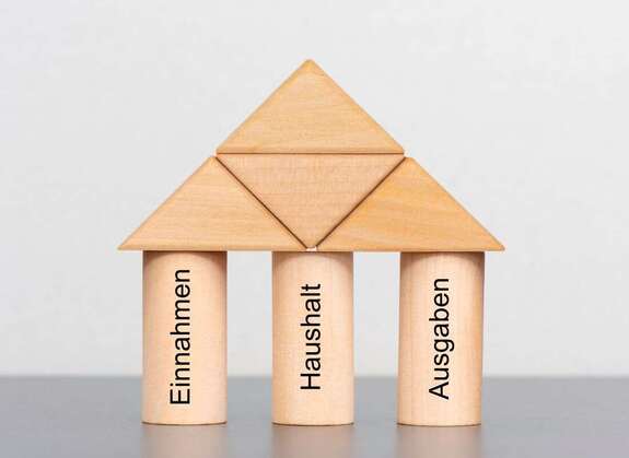 Holzwürfel als Haus aufgebaut mit den Schriftzügen Haushalt, Einnahmen, Ausgaben