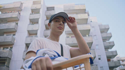 Filmszene Masel Tov Cocktail. Junger Mann steht vor einem Gebäudekomplex.