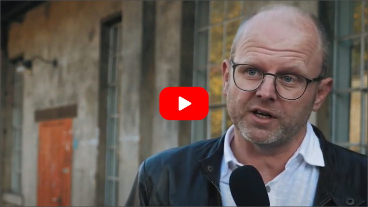 Foto: Videovorschaubild, Matthias Hoffmann mit weißem Hemd und schwarzer Jacke, im Hintergrund einstöckige Häuser aus Stein